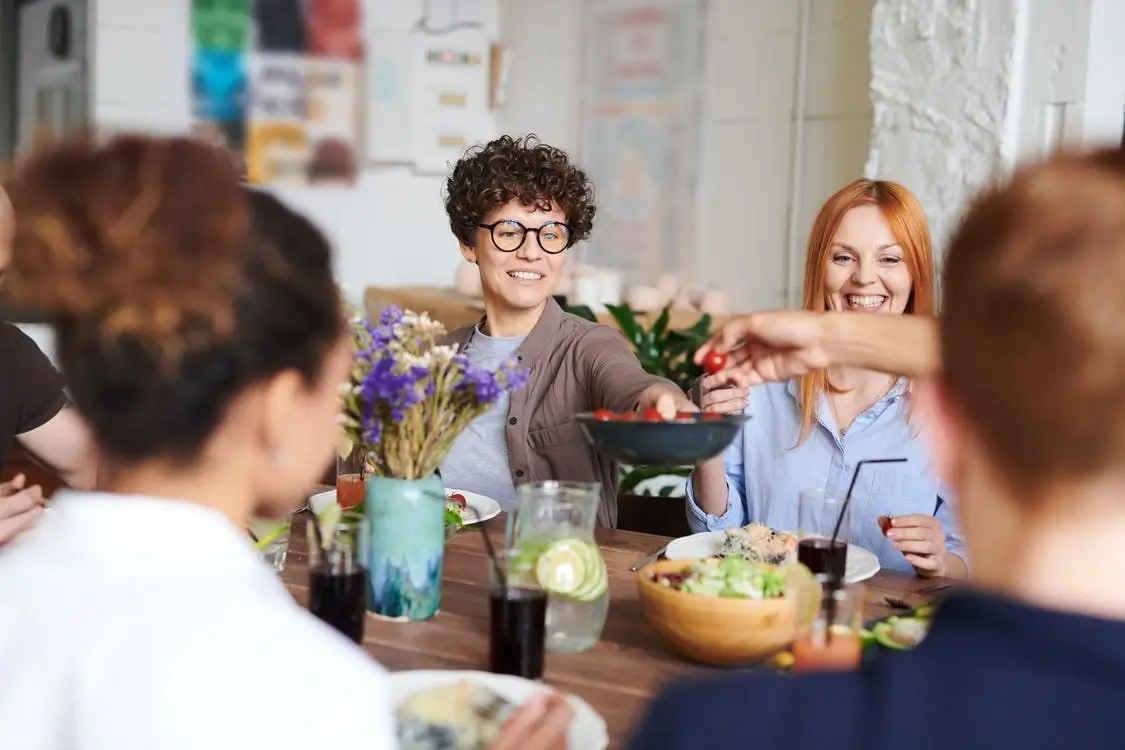 Menschen sitzen an einem Tisch. Frau lächelt und hält Schale mit Essen in die Mitte des Tisches.