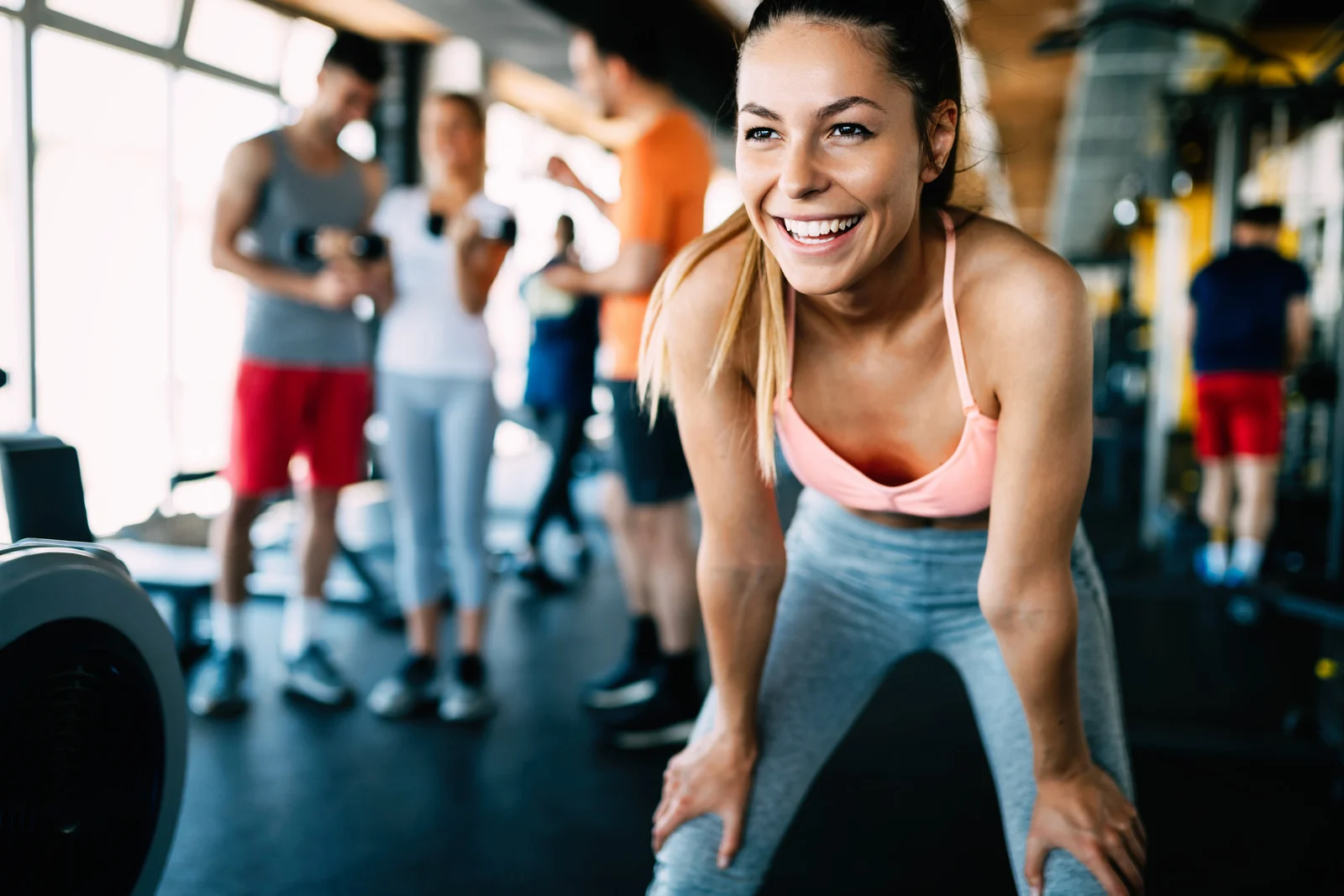 Lachende Frau in Sportkleidung macht eine Pause im Fitnessstudio; im Hintergrund trainieren weitere Personen an Geräten.