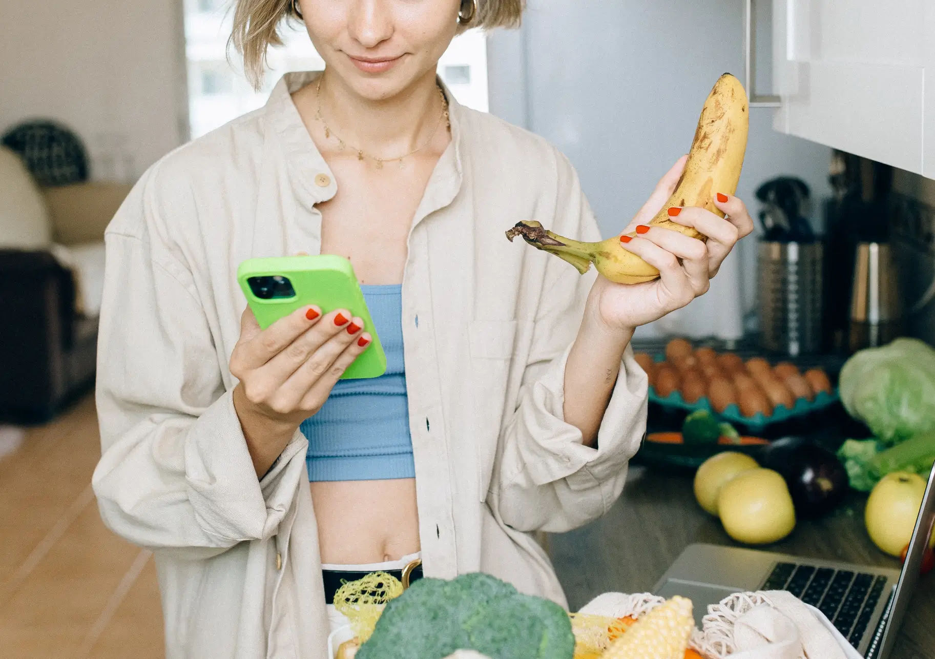 Frau steht in der Küche und hält eine Banane in der linken und ein Handy in der rechten Hand.