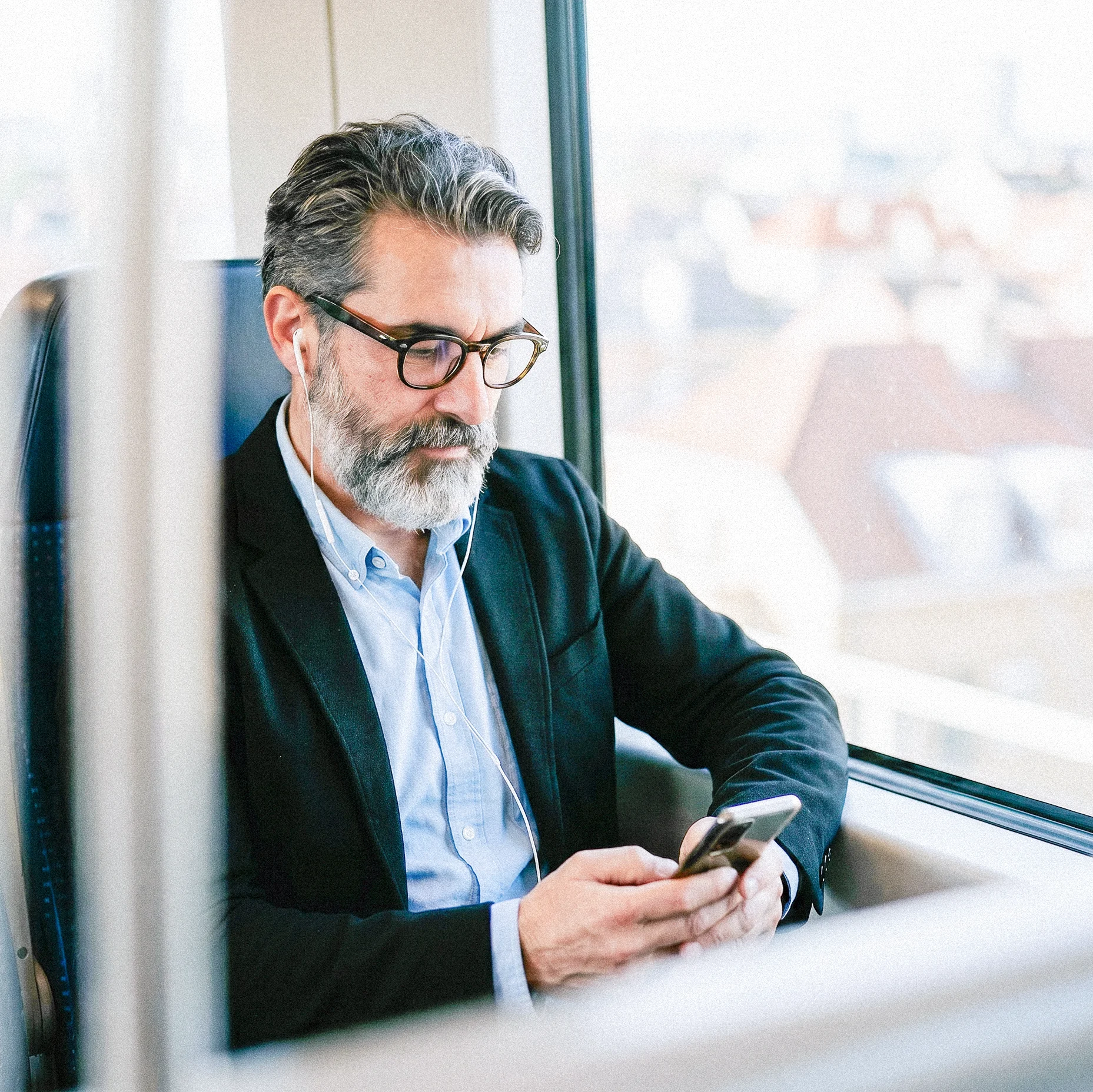 Mann mit Brille und Kopfhörern sitzt im Zug am Fenster und nutzt sein Smartphone; draußen ist eine unscharfe Stadtlandschaft zu sehen.