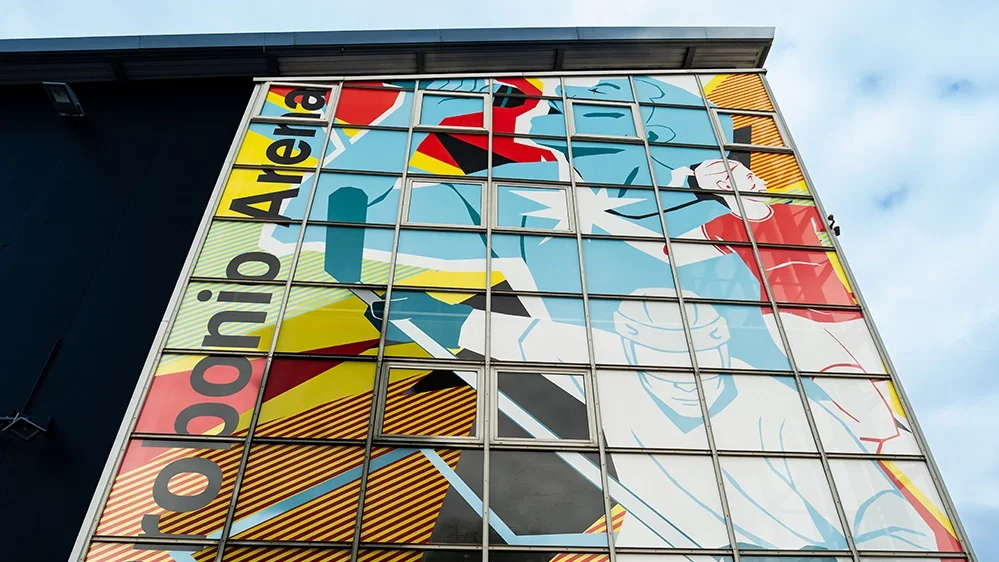 Neue Glasfassade der Probonio Arena eingeweiht Außenansicht eines Gebäudes mit großer Glasfassade und farbigem Motiv; links steht vertikal der Schriftzug „Probonio Arena“.