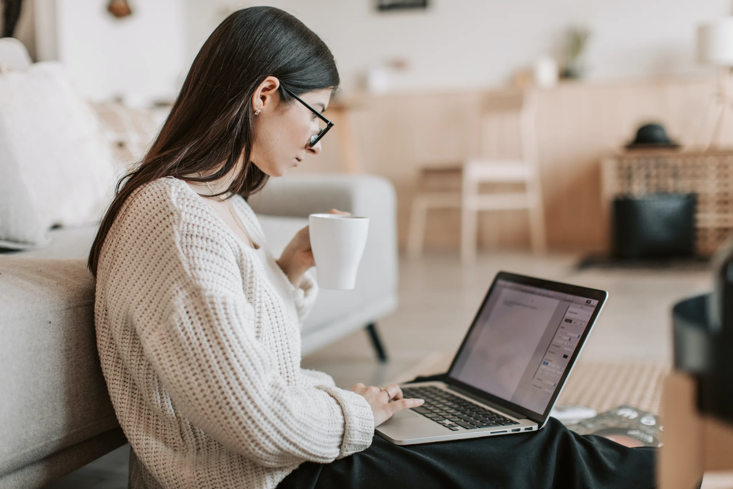 Frau sitzt am Boden mit Laptop auf dem Schoß und Kaffeetasse in der Hand.
