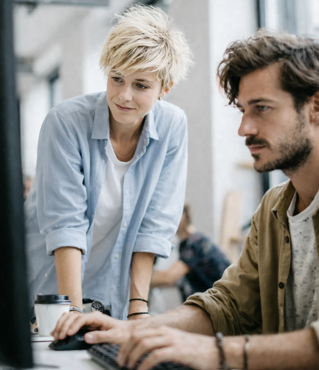 Ein Mann sitzt an seinem Arbeitsplatz und schaut in seinen Computer. Eine Frau steht daneben und schaut auch auf den Computer