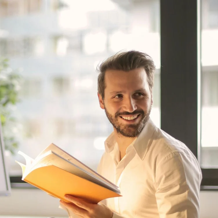 Bonuszahlung: Tipps zur Abrechnung und Versteuerung Ein Mann sitzt an einem Schreibtisch im Büro und liest ein Buch. Neben ihm stehen ein Monitor und eine Schreibtischlampe, im Hintergrund sind große Fenster mit Blick auf ein Gebäude zu sehen.