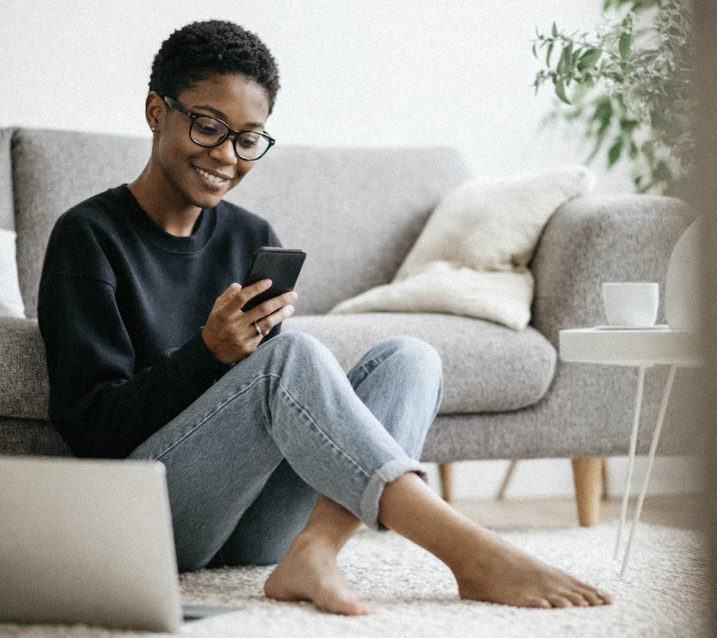 Eine Frau sitzt am Boden und hält ihr Handy in der Hand. Ihr Laptop steht am Boden neben ihr