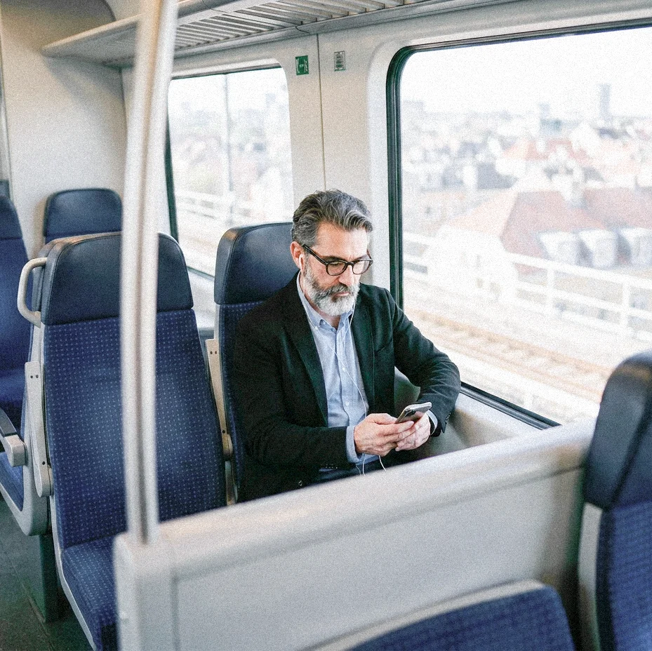 Mann sitzt allein in einem Zug am Fenster und blickt auf sein Smartphone; durch die Scheibe ist eine Stadtansicht zu sehen.