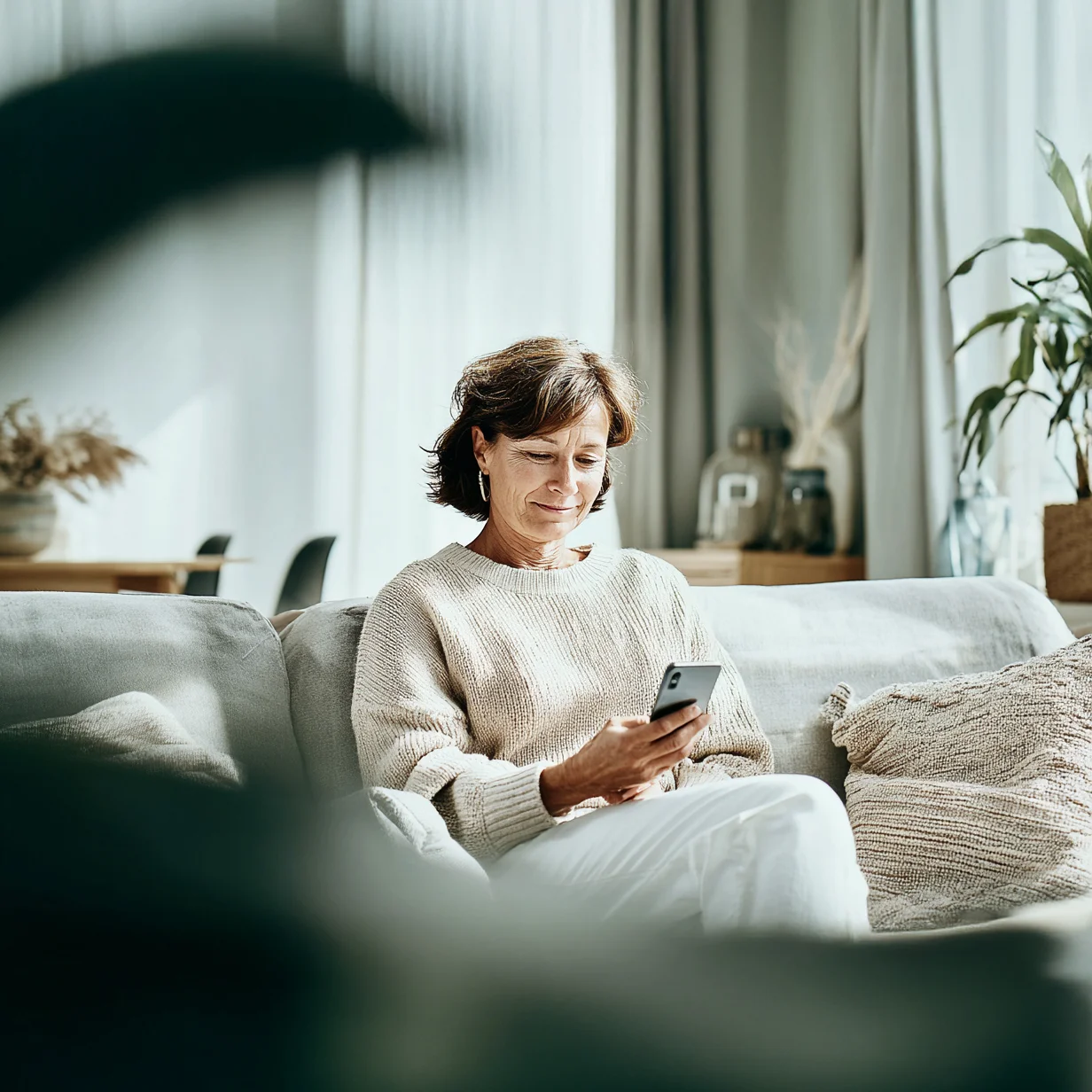 Benefit Lexikon betriebliche Altersvorsorge Ältere Frau sitzt mit dem Smartphone in der Hand auf dem Sofa und lächelt