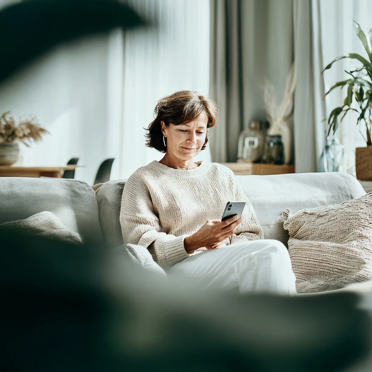 Ältere Frau sitzt mit dem Smartphone in der Hand auf dem Sofa und lächelt