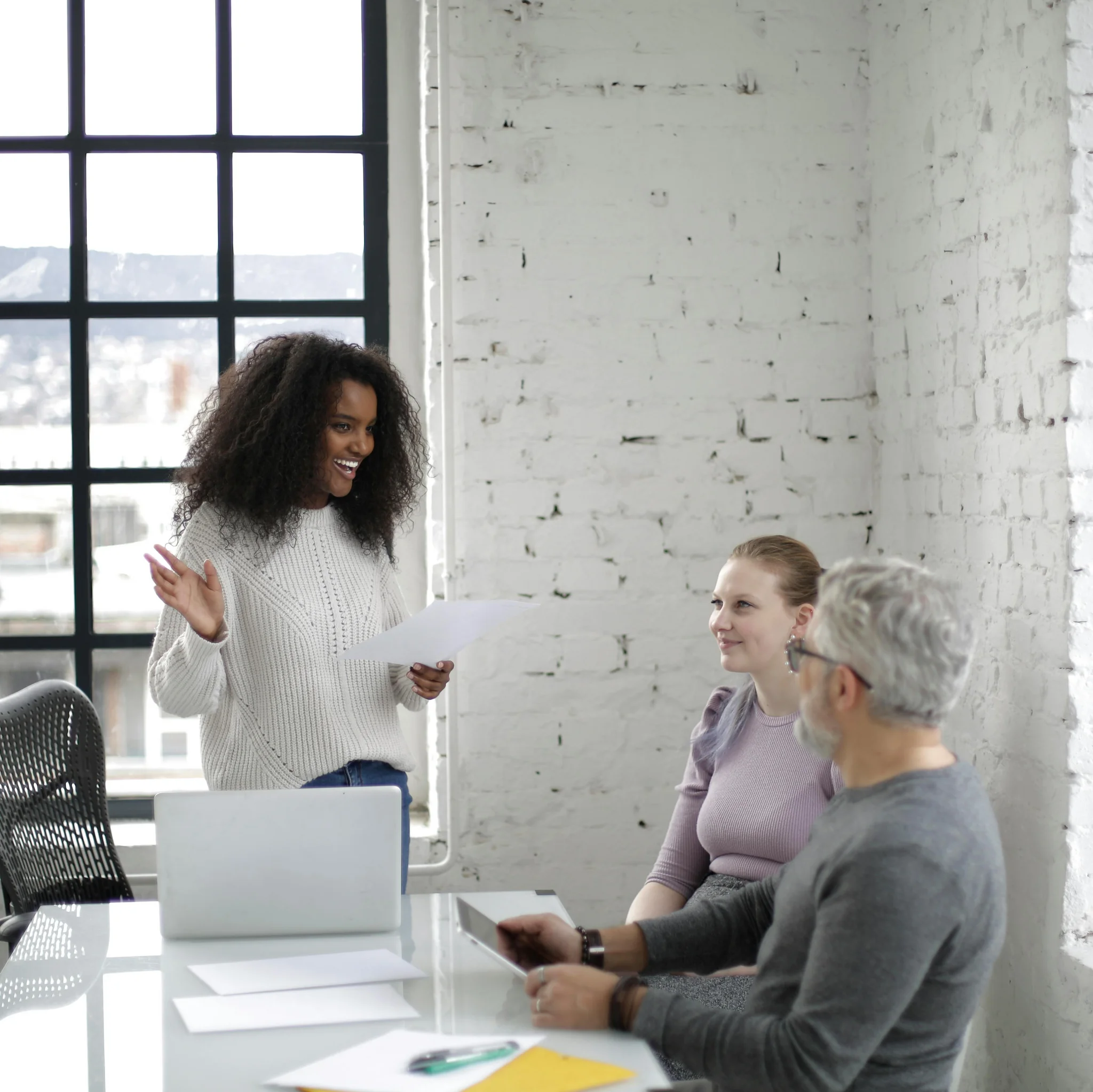 Eine Frau präsentiert ein Dokument in einem modernen Büro, während zwei Kolleg:innen am Tisch sitzen und zuhören. Auf dem Tisch liegen Laptop, Tablet und Unterlagen.