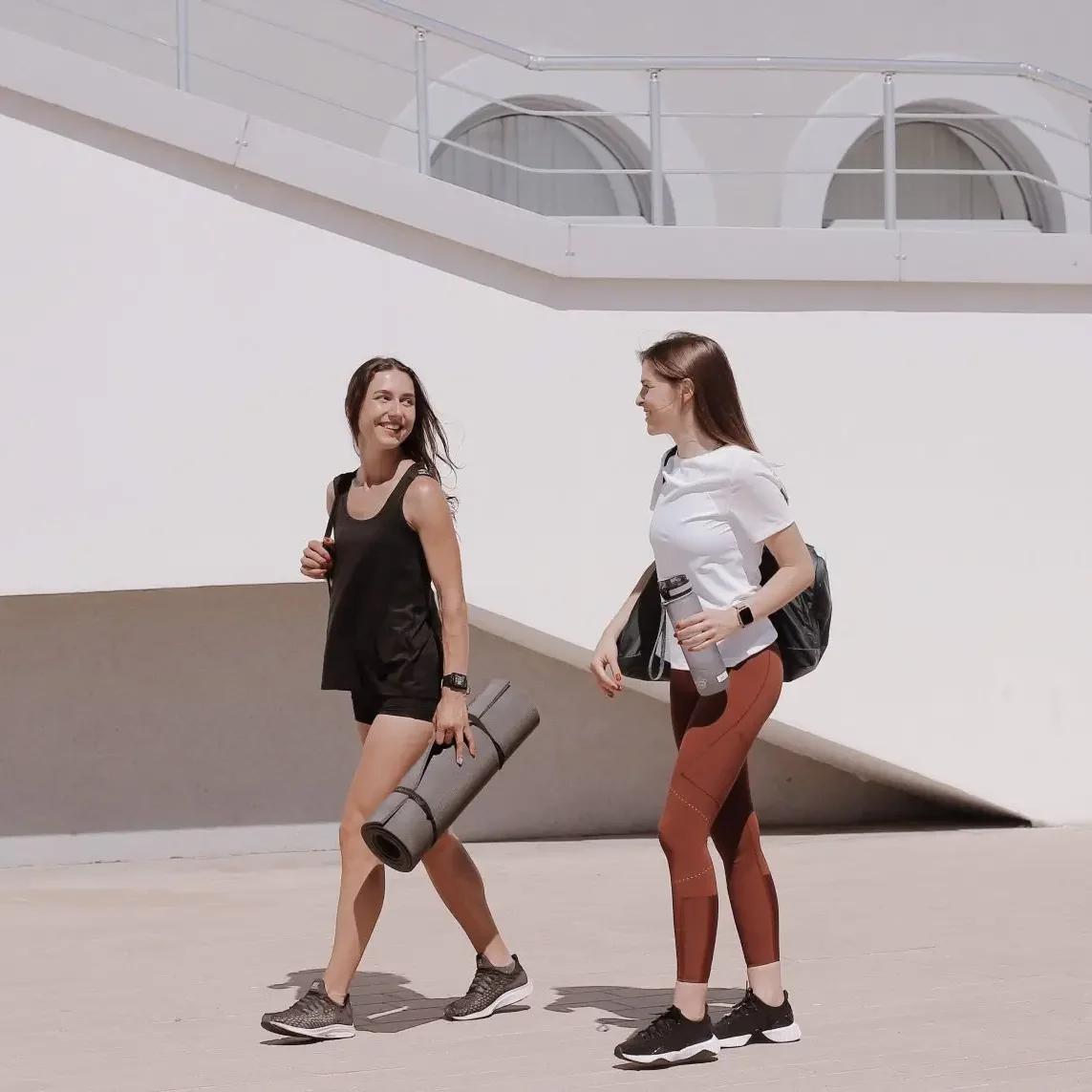 2 Frauen in Sportkleidung gehen vor einem Gebäude entlang. Eine Frau hält eine Yogamatte in der Hand. Die andere eine Trinkflasche und eine Sporttasche.