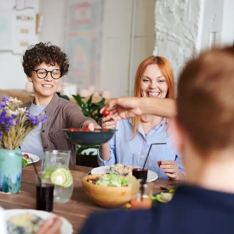 Menschen sitzen an einem Tisch. Frau lächelt und hält Schale mit Essen in die Mitte des Tisches.