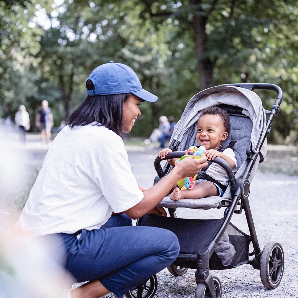 Eine Person kniet vor einem Kinderwagen und reicht einem lächelnden Baby ein Spielzeug; im Hintergrund ist ein Parkweg mit Bäumen zu sehen.