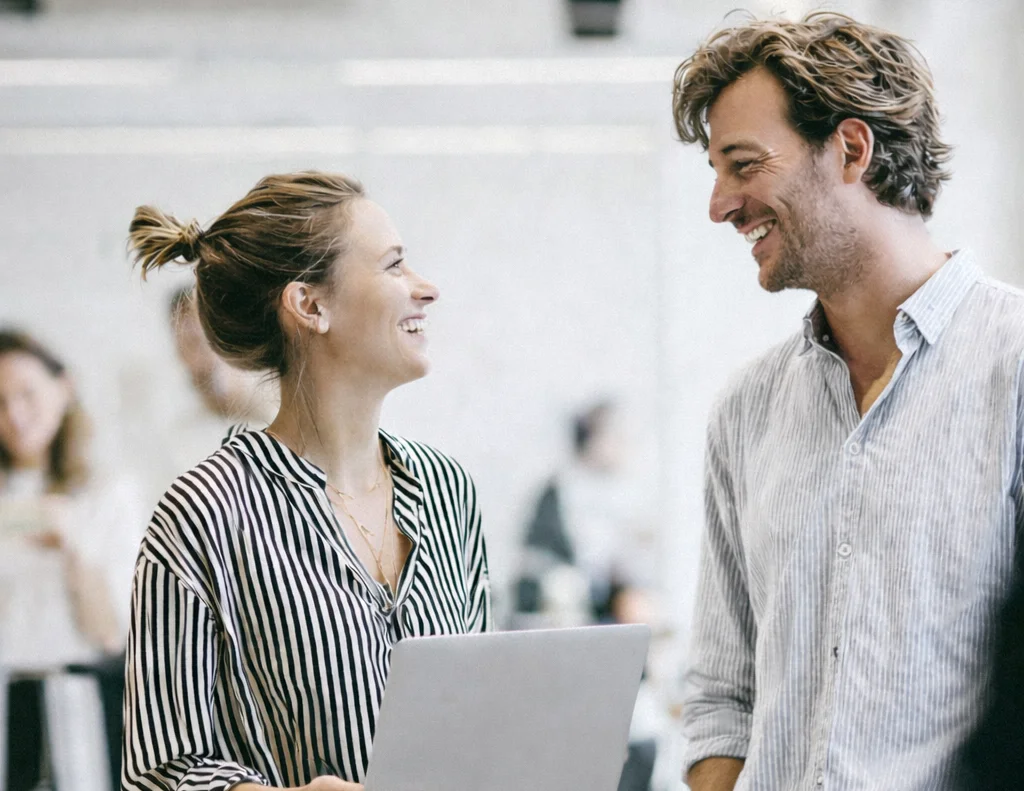 Zwei Personen lachen zusammen im Büro. Sie hält ihren Laptop in der Hand