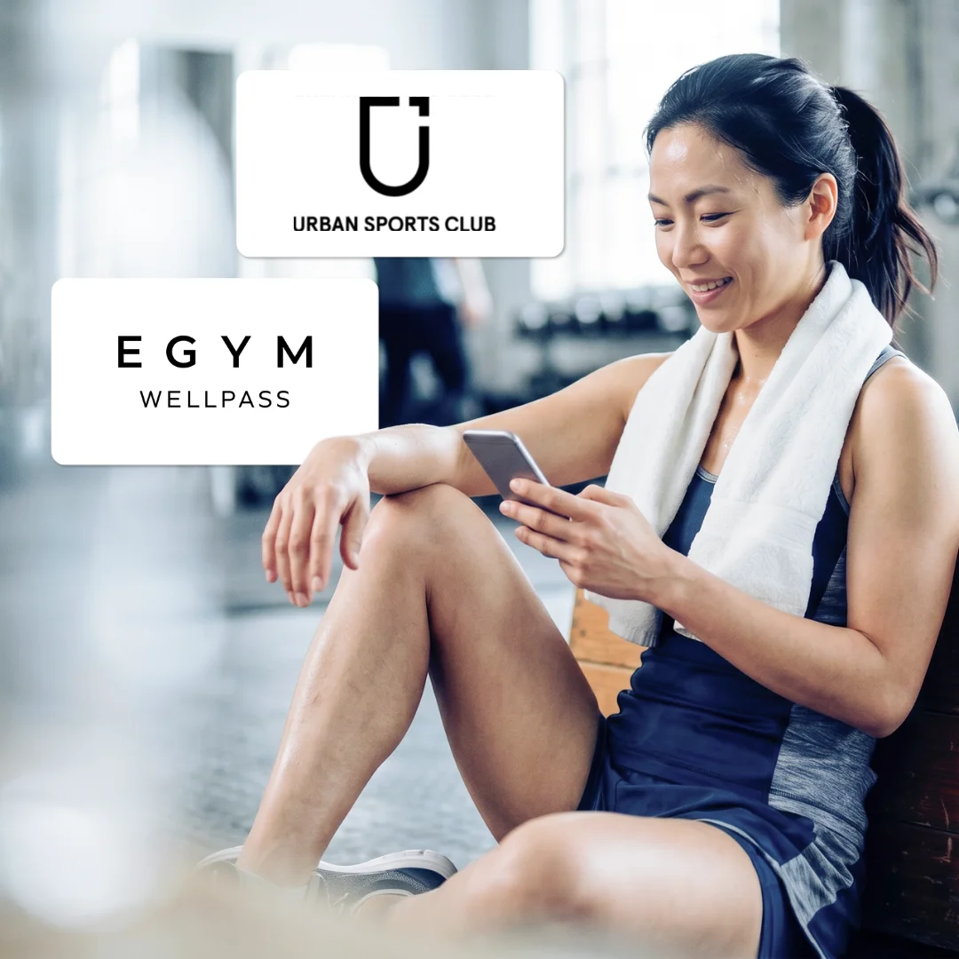Sportliche Frau sitzt im Fitnessstudio mit Handtuch um den Hals und schaut auf ihr Smartphone; eingeblendete Logos „Urban Sports Club“ und „EGYM Wellpass“.
