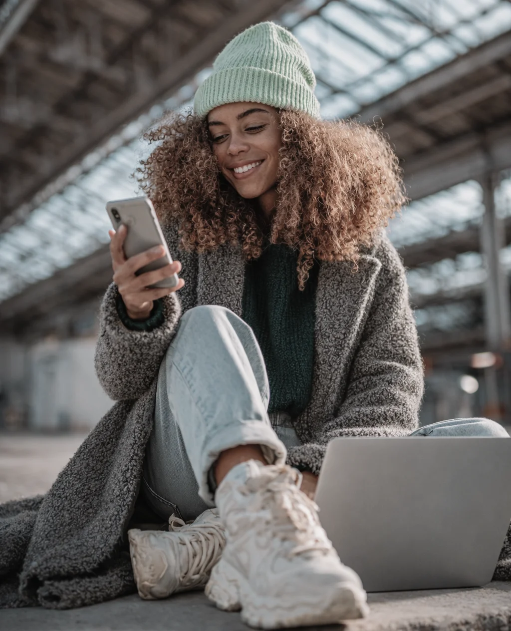 Eine Frau sitzt am Bahnhof und lacht in ihr Handy. Vor ihr steht ein Laptop.