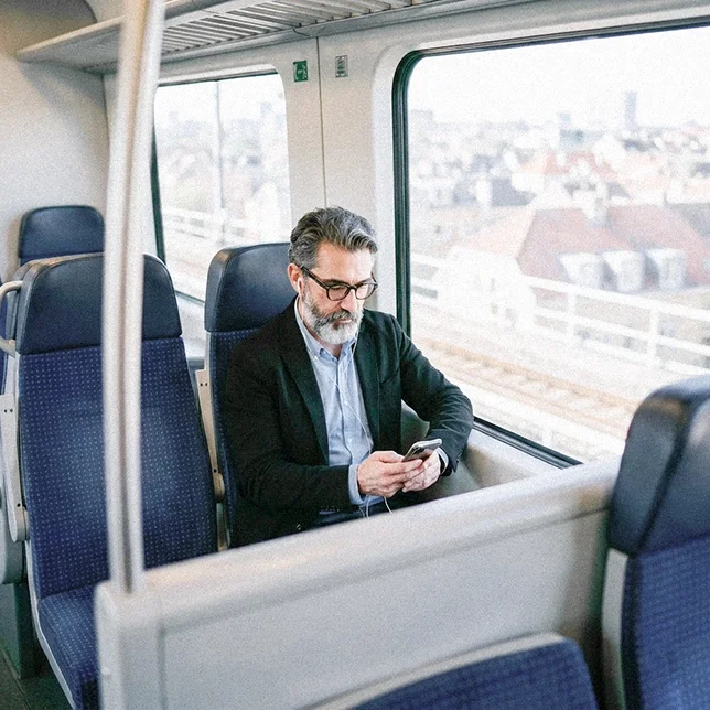 Mann sitzt allein in einem Zugabteil am Fenster und nutzt ein Smartphone; durch das Fenster ist eine Stadtansicht zu sehen.