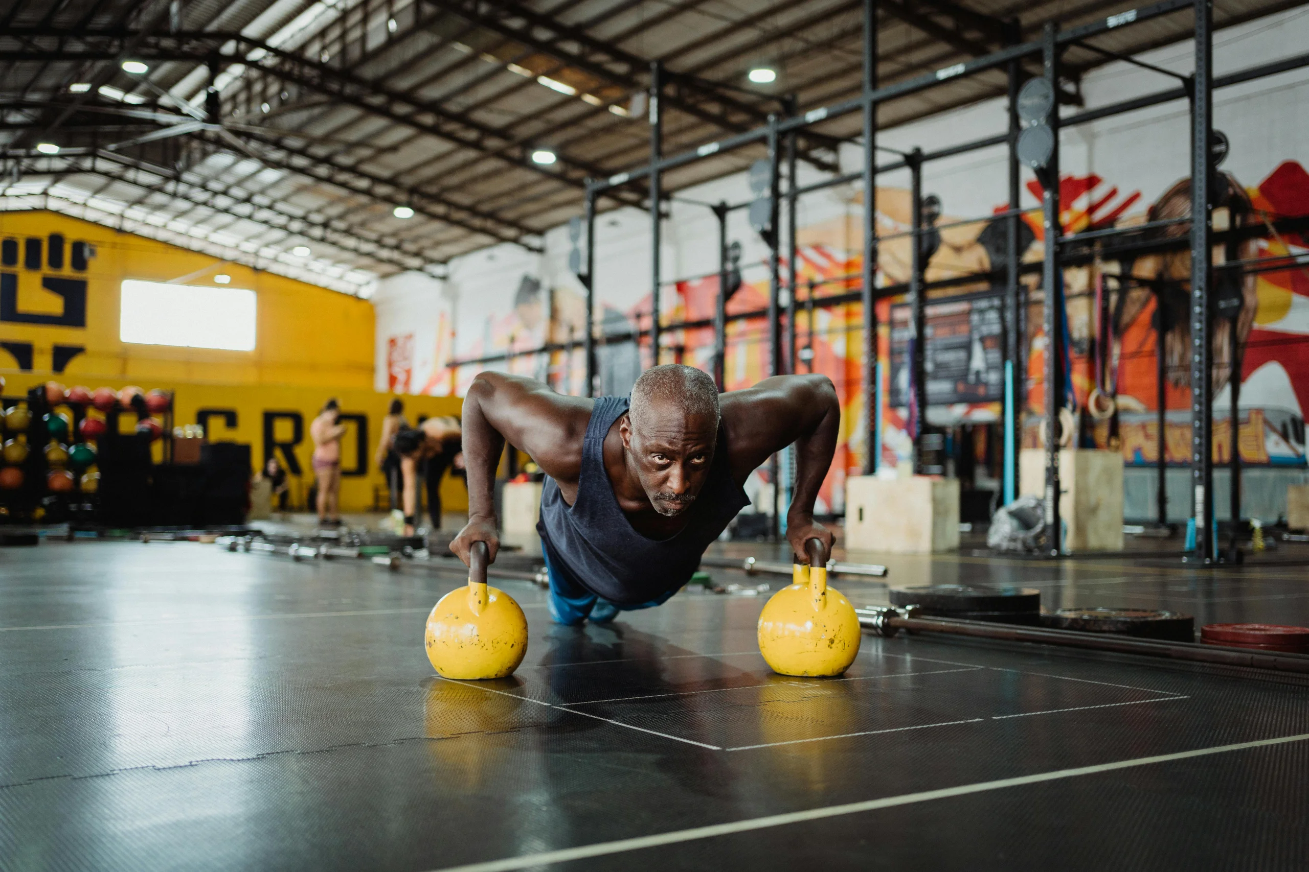 Mann macht Liegestütze auf zwei Kettlebells in einer großen Trainingshalle; im Hintergrund sind weitere Trainierende und Fitnessgeräte zu sehen.