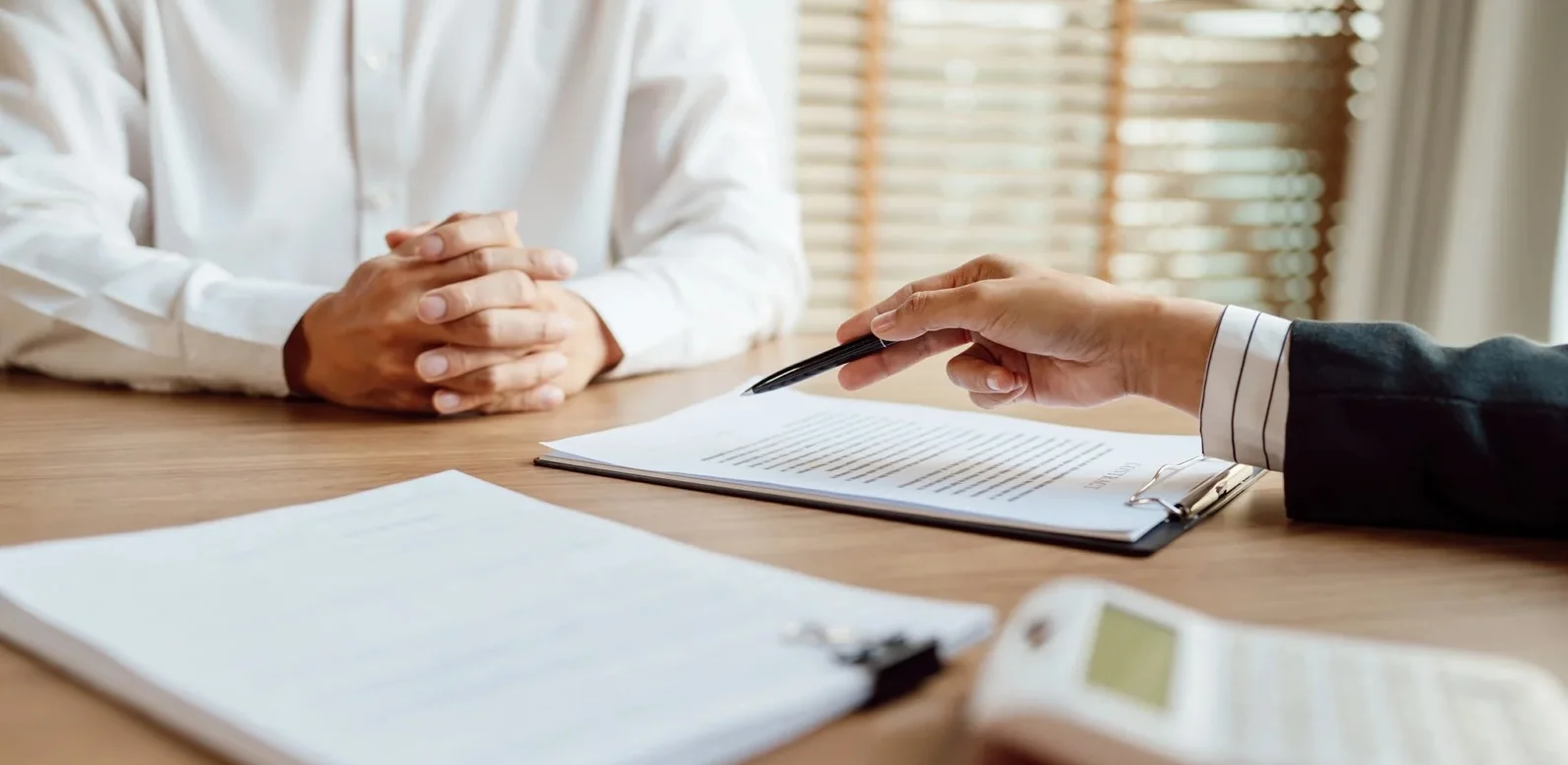 Auf einem Tisch liegen Unterlagen. Ein Oberkörper mit verschränkten Armen und eine Hand, welche mit einem Kugelschreiber etwas auf einem Papier zeigt.