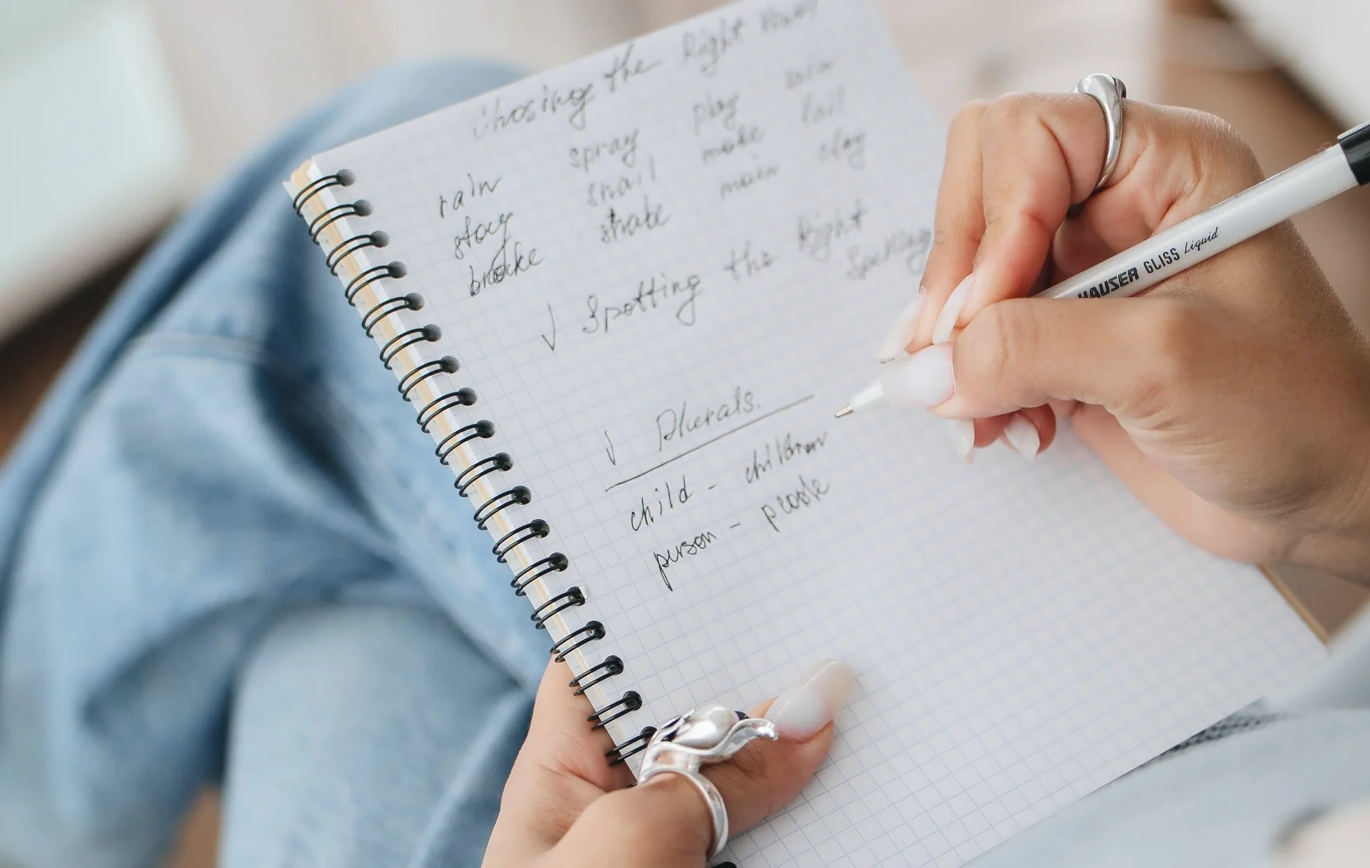 Nahaufnahme einer Hand, die mit einem Stift Notizen in ein kariertes Spiralheft schreibt; im Hintergrund ist unscharf eine Jeans zu sehen.