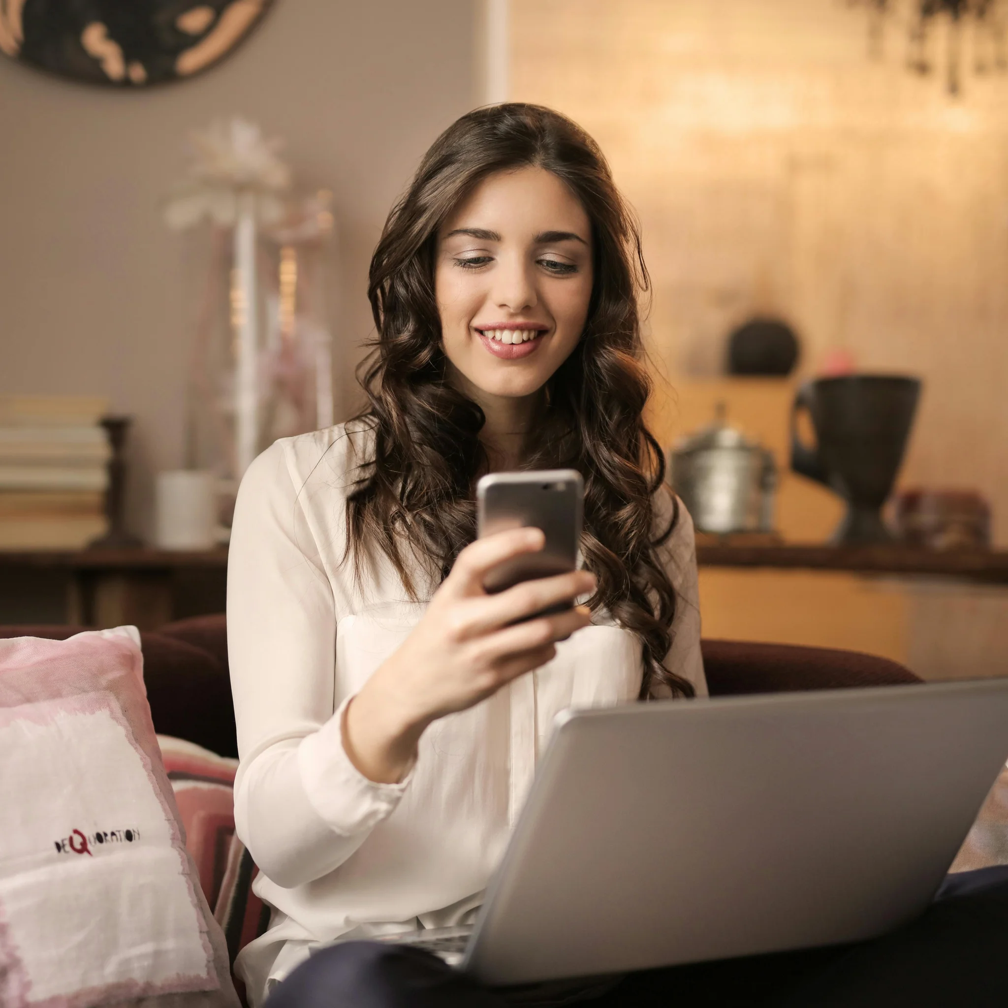 Frau sitz auf einem Sofa. Sie hat einen Laptop auf ihrem Schoß und schaut lächelnd auf ihr Handy.