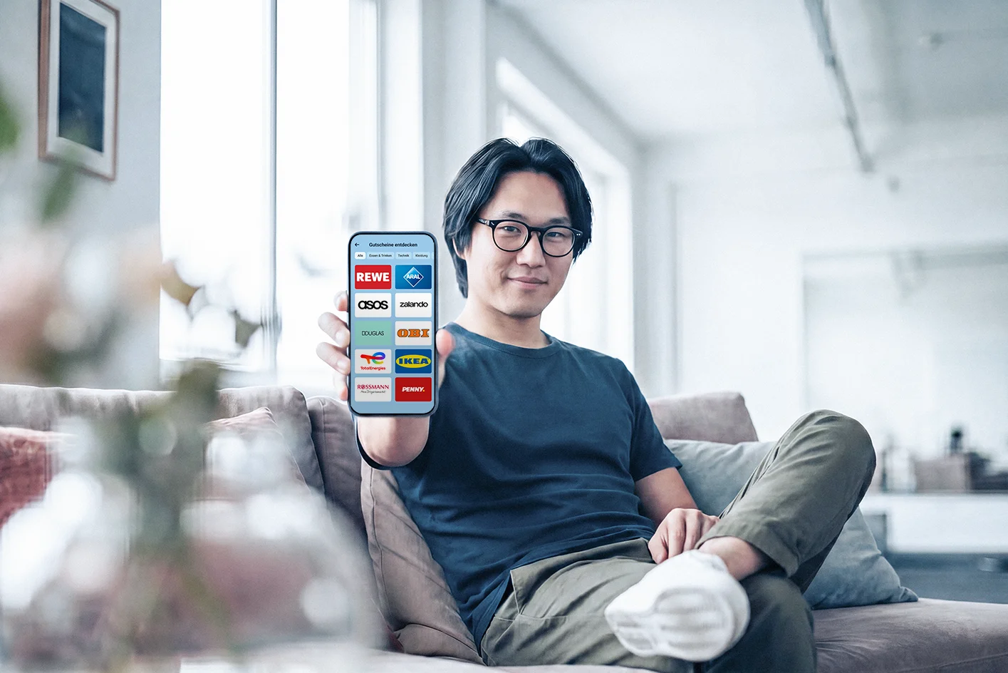 Mann sitzt auf dem Sofa und hält ein Smartphone mit einer Übersicht digitaler Gutscheine in die Kamera.